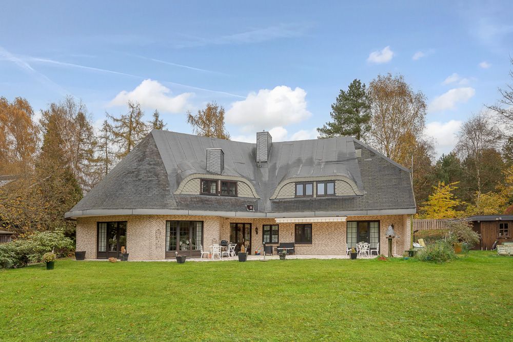 Großes Haus mit dunklem Dach, vielen Fenstern und Terrasse, umgeben von Garten und Bäumen.