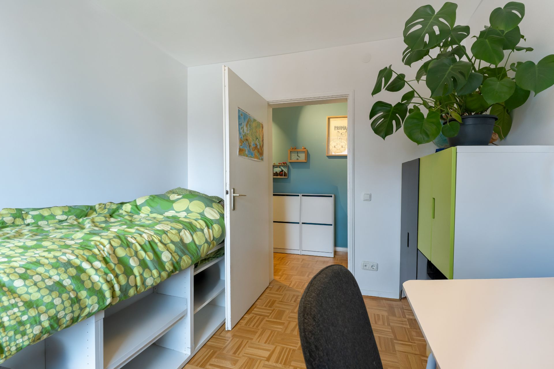 Kleines Zimmer mit Einzelbett, grünem Bettbezug, Schreibtisch, Stuhl und großer Zimmerpflanze.