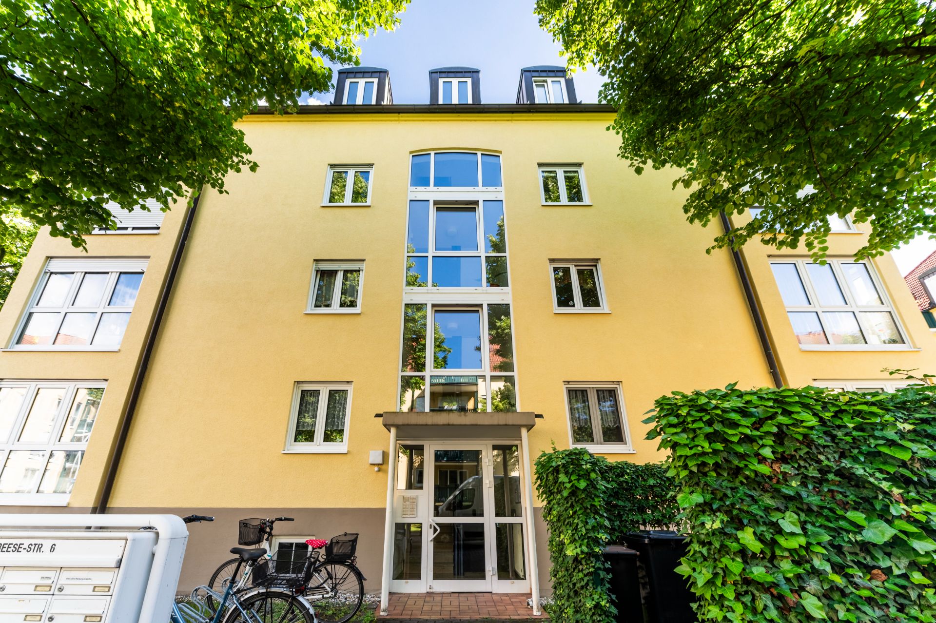 Gelbes Wohnhaus mit vielen Fenstern, Eingangstür und zwei Fahrrädern davor, umgeben von Bäumen.