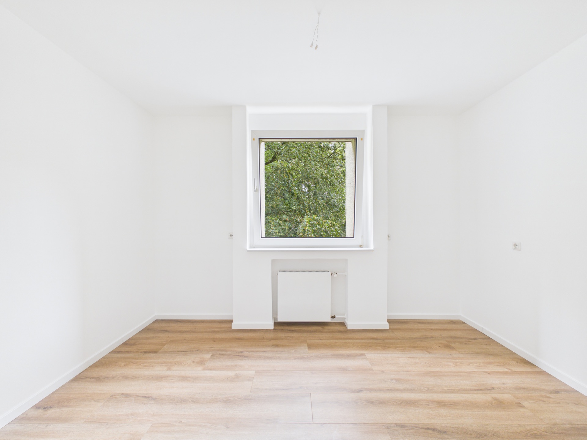 Leeres Zimmer mit weißem Wänden, Holzfußboden und einem Fenster mit Blick ins Grüne.