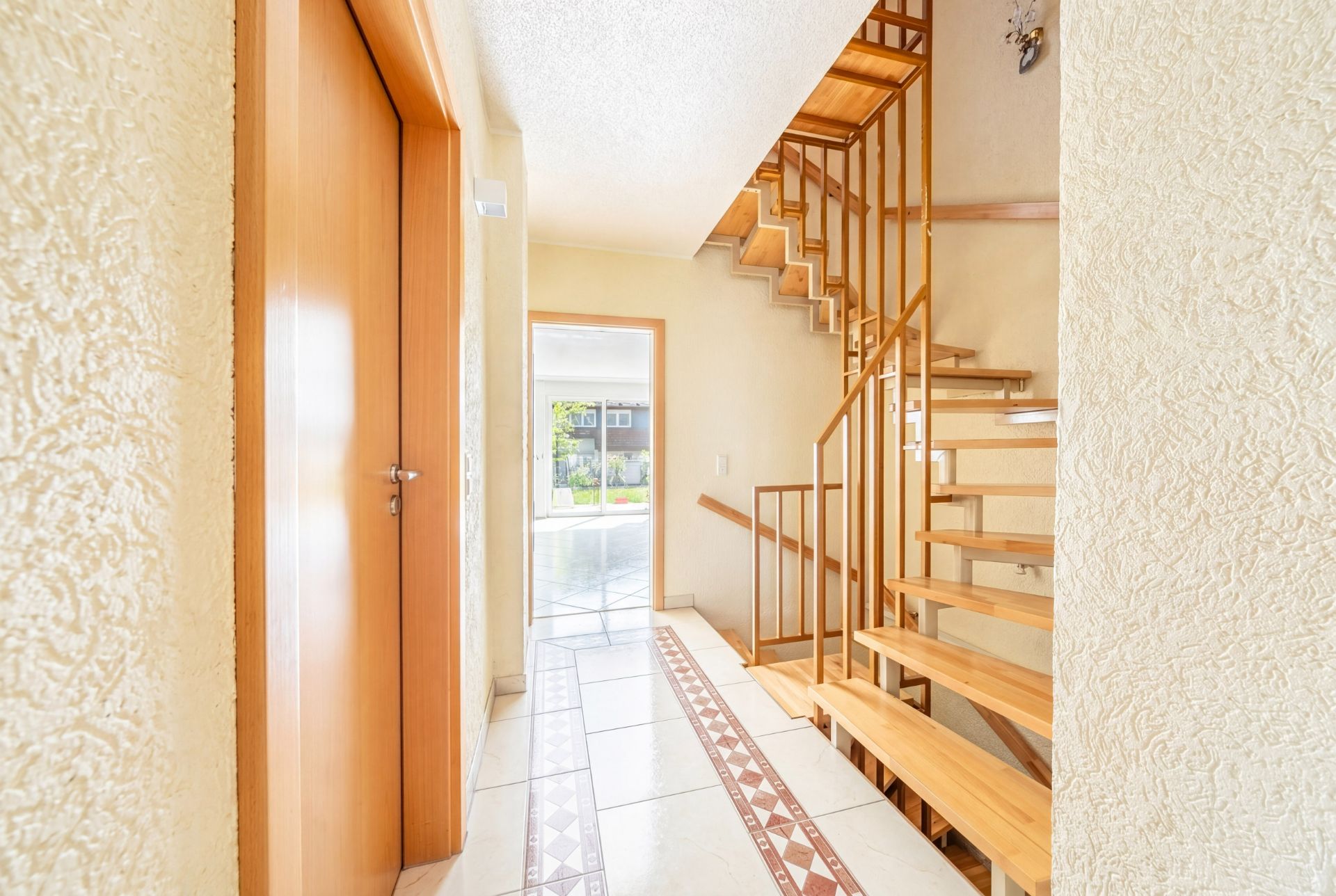 Heller Flur mit Holztür, gemusterten Fliesen und offener Holztreppe.