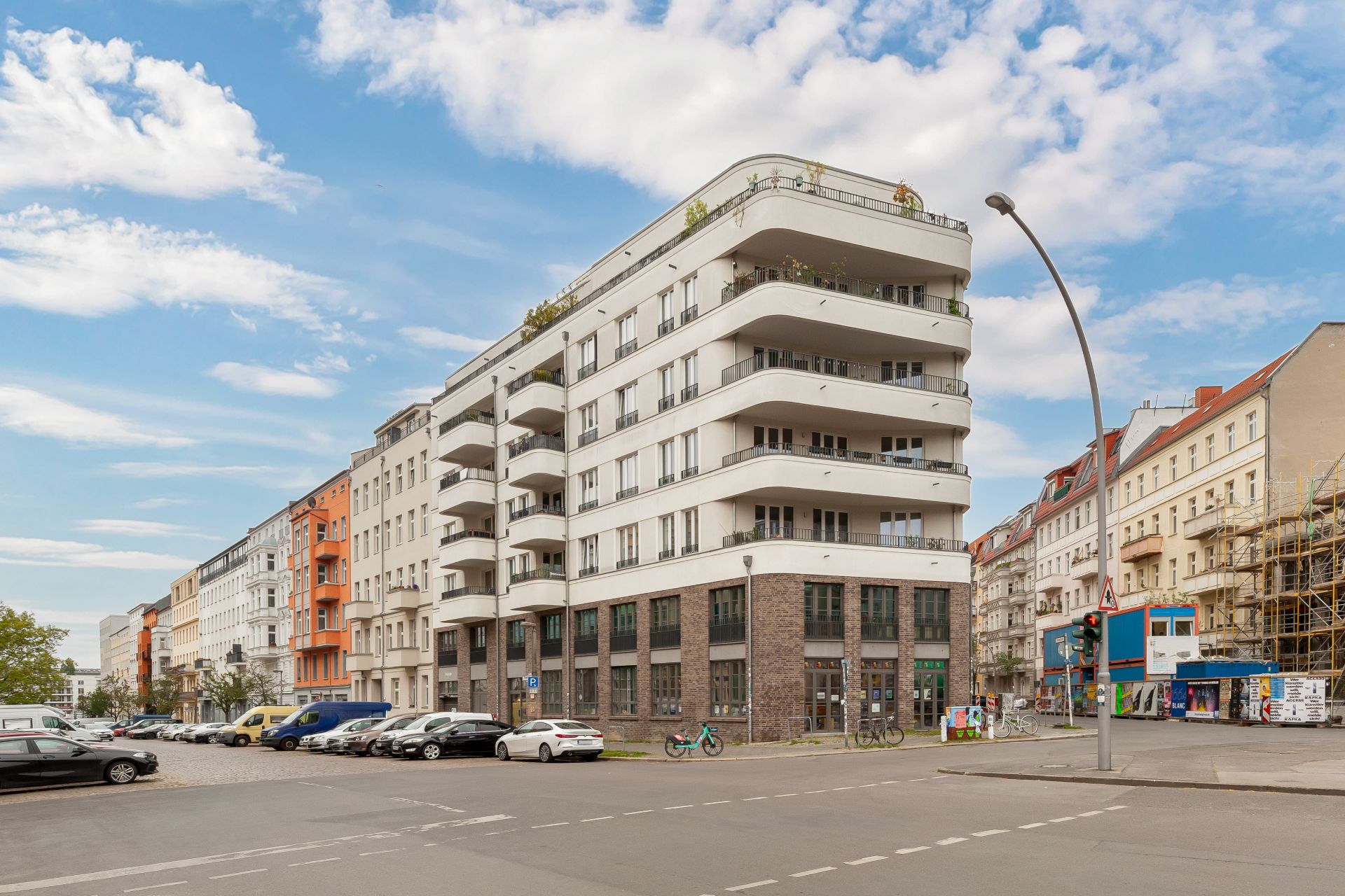 Modernes Wohngebäude mit abgerundeten Balkonen an einer Straßenecke, geparkte Autos und Fahrräder davor.