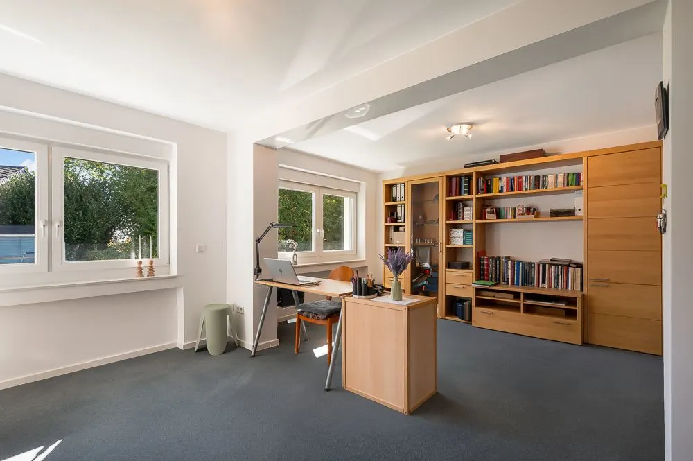 Helles Arbeitszimmer mit Schreibtisch, Bücherregal und zwei großen Fenstern.