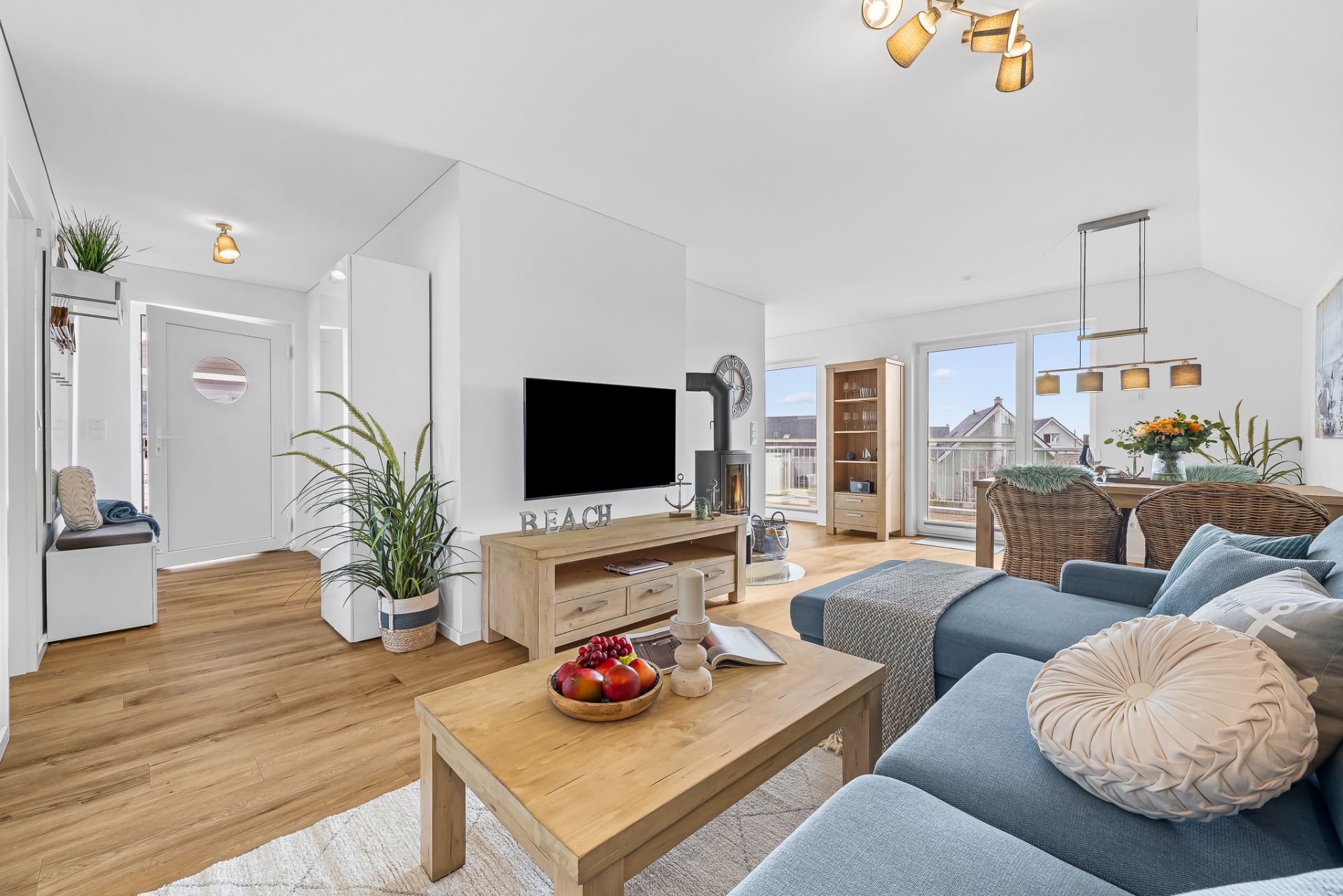 Helles Wohnzimmer mit Sofa, Couchtisch, Fernseher und Esstisch, große Fenster und Holzboden.