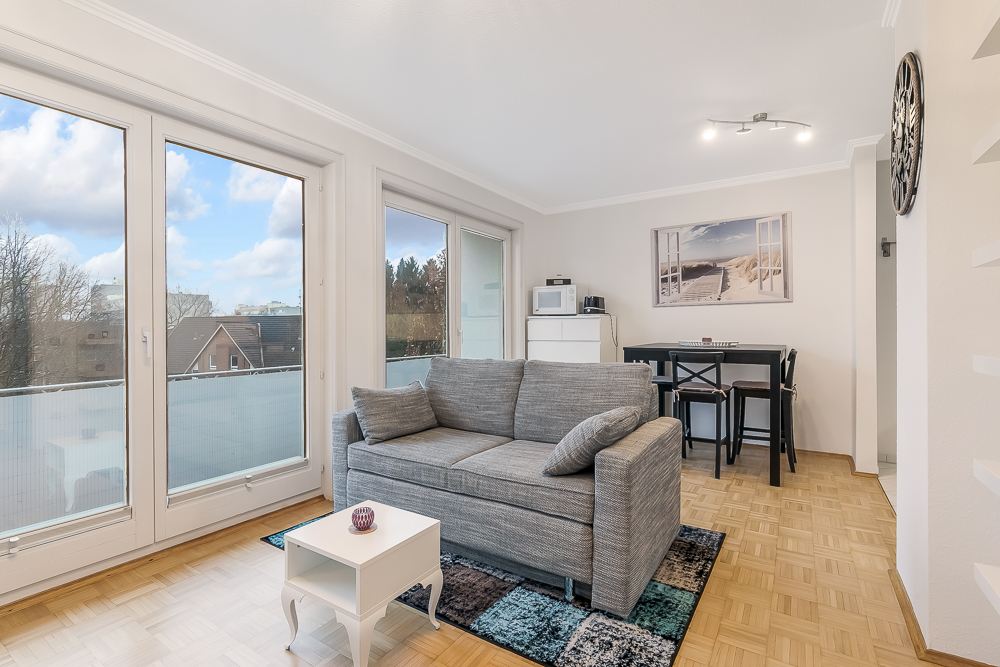 Helles Wohnzimmer mit grauem Sofa, kleinem weißen Tisch und Essbereich vor großen Fenstern.
