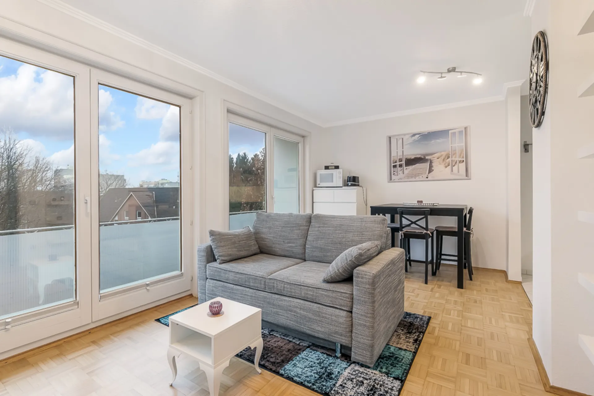 Helles Wohnzimmer mit grauem Sofa, kleinem weißen Tisch und Essbereich vor großen Fenstern.