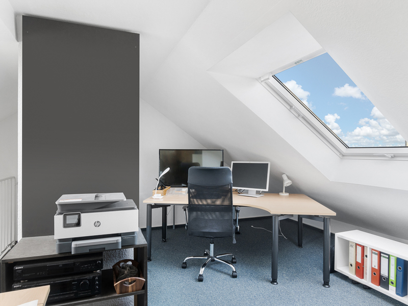 Helles Arbeitszimmer mit Schreibtisch, Stuhl, Monitoren und Dachfenster in einem Dachgeschoss.