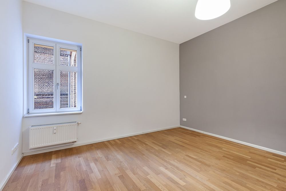 Leerer Raum mit Holzboden, weißem Fenster und grauer Akzentwand.