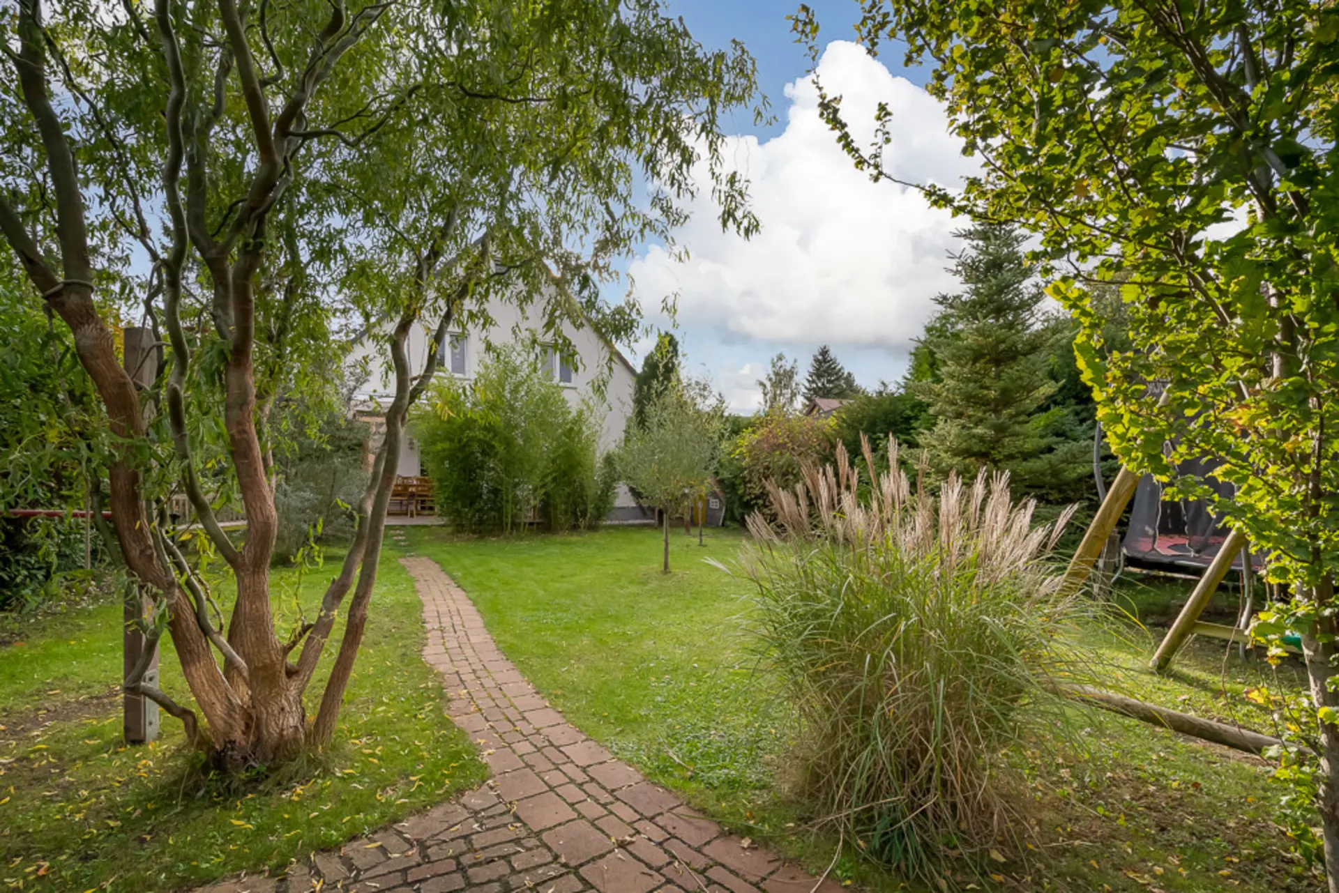 Garten mit Rasen, Bäumen, Sträuchern und gepflastertem Weg zu einem Wohnhaus.