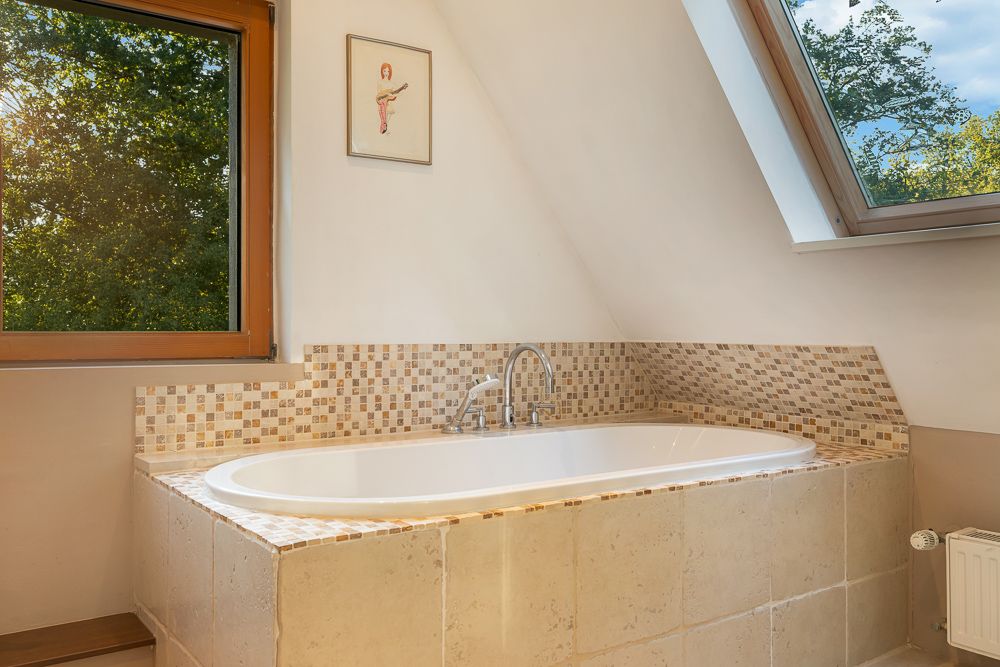 Badewanne unter Dachschräge mit Mosaikfliesen, großem Fenster und Blick ins Grüne.