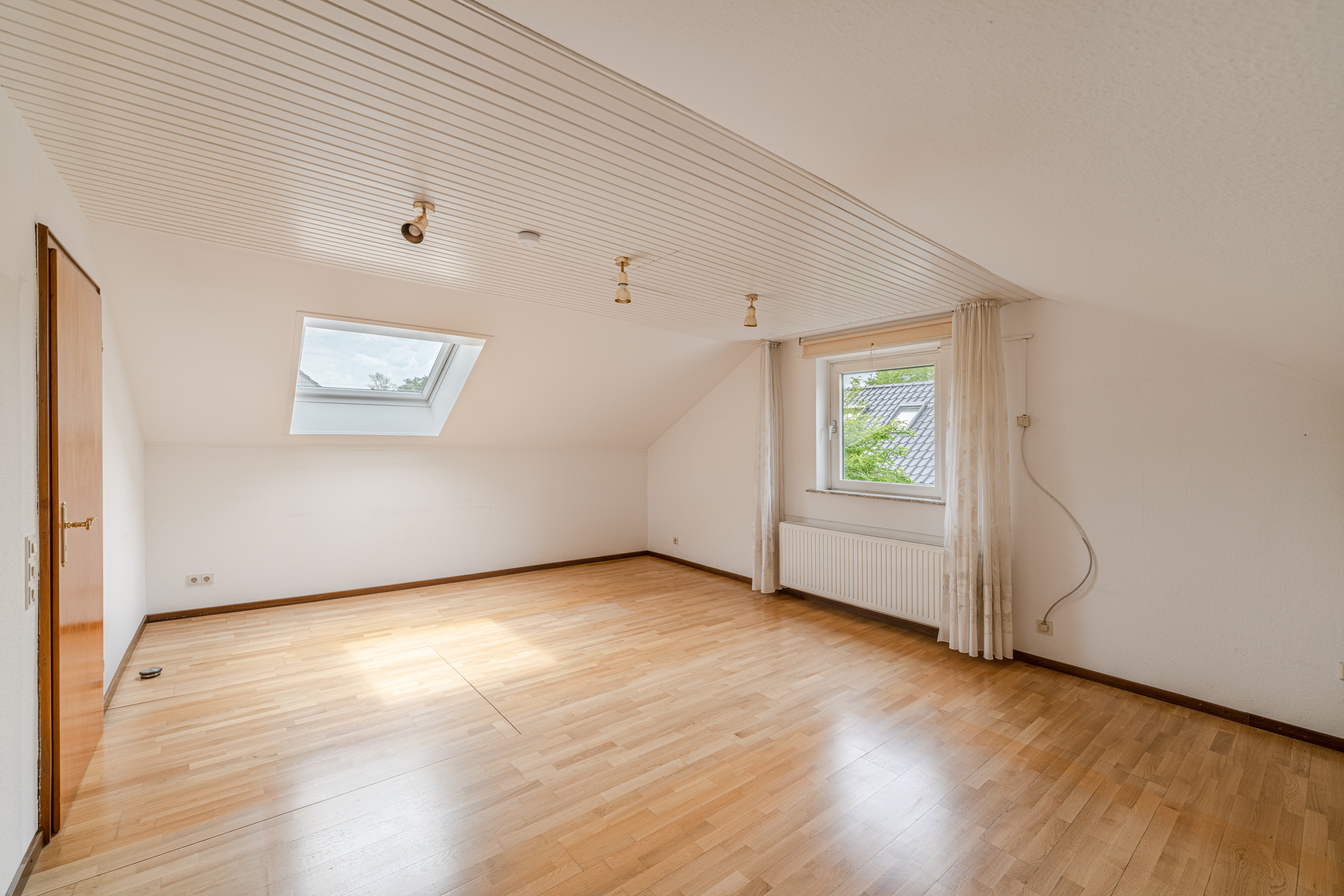 Helles Zimmer mit Holzfußboden, Dachfenster und normalem Fenster, weiße Wände und Decke.