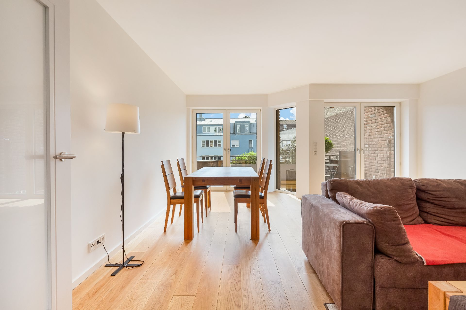 Helles Wohnzimmer mit Esstisch, vier Stühlen, Stehlampe und braunem Sofa vor großen Fenstern.