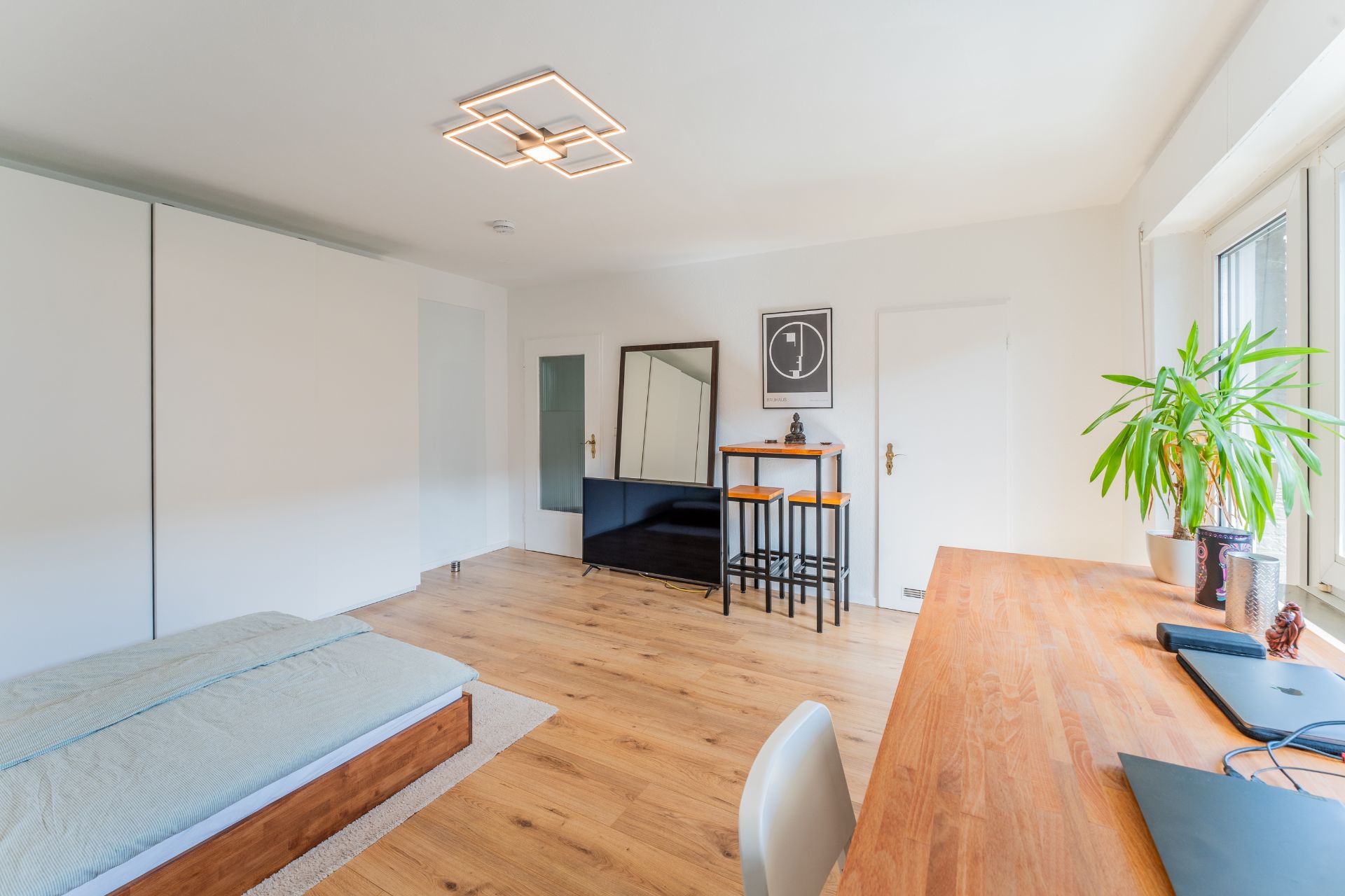 Helles Zimmer mit Bett, Schreibtisch, Pflanzen, Esstisch und großem Spiegel auf Holzfußboden.
