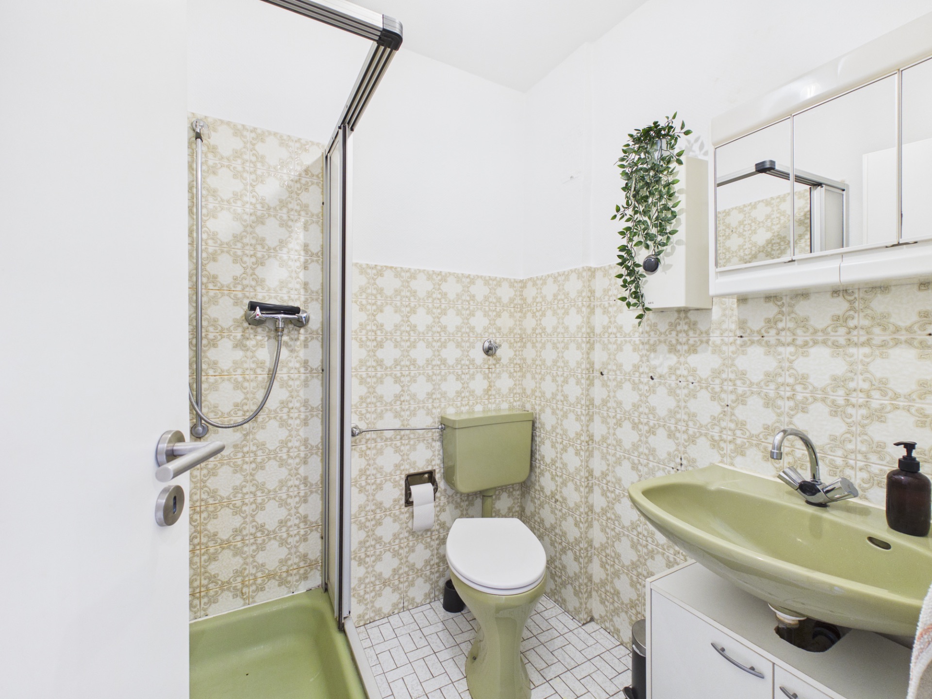 Kleines Badezimmer mit grüner Dusche, Toilette und Waschbecken sowie gemusterten Fliesen.