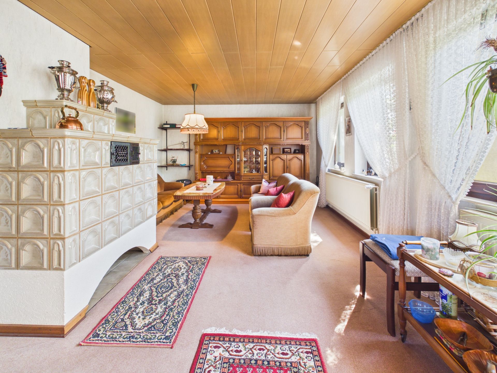Helles Wohnzimmer mit Kachelofen, Teppichen, Sofa, Holzschränken und großen Fenstern mit Gardinen.