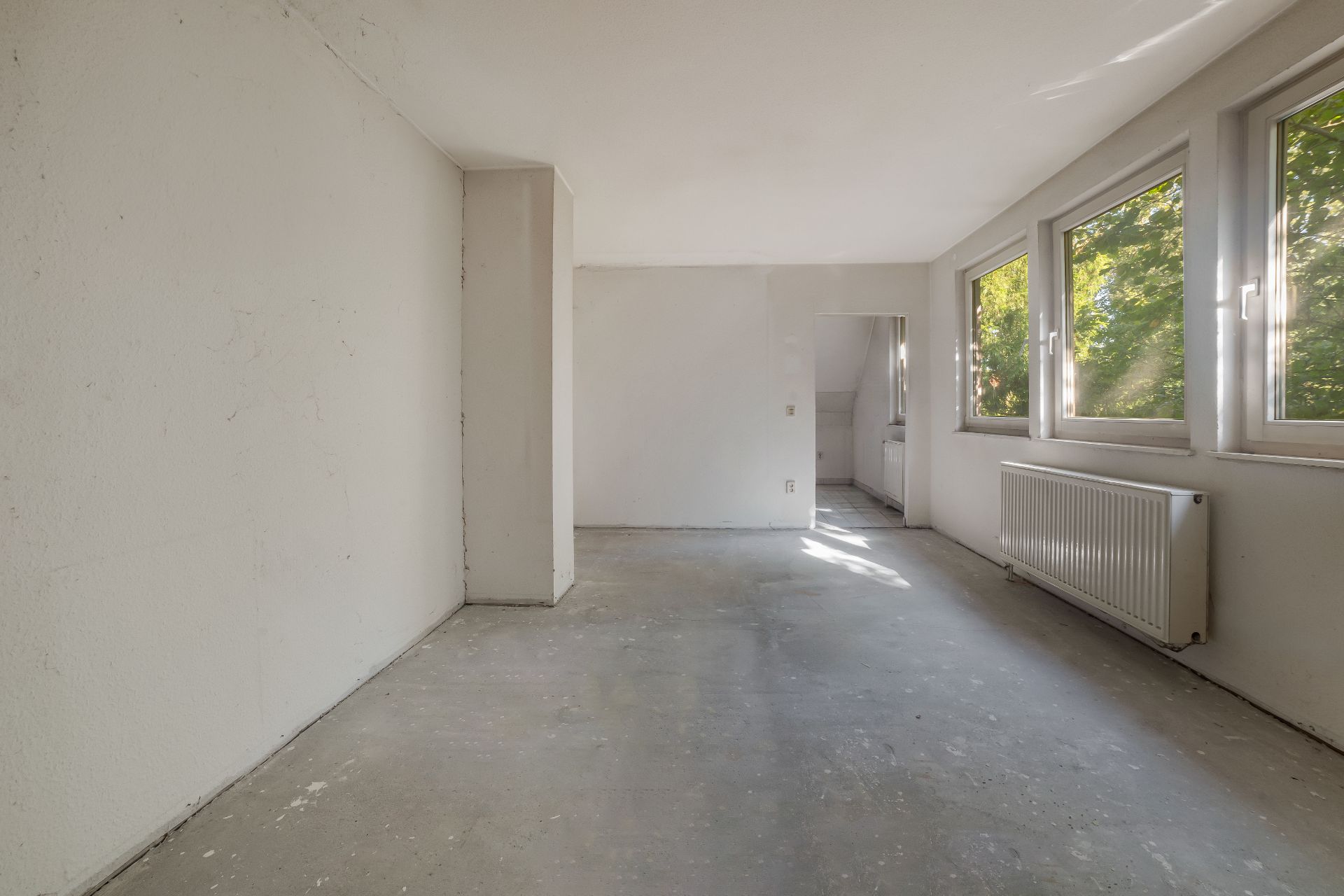 Leerer Raum mit weißen Wänden, großen Fenstern und Heizkörper, Blick auf Bäume draußen.