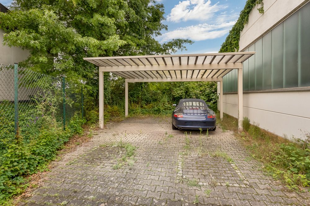 Überdachter Stellplatz mit einem geparkten Auto, umgeben von Pflanzen und einem Zaun.