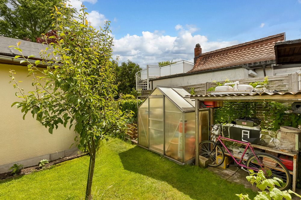 Garten mit kleinem Gewächshaus, Baum und überdachtem Abstellbereich für Fahrräder und Werkzeuge.