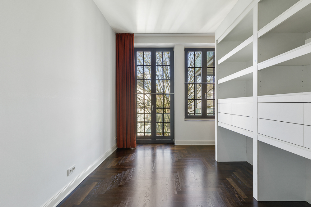 Heller Raum mit dunklem Holzboden, Regalwand und Fensterfront mit rotem Vorhang.