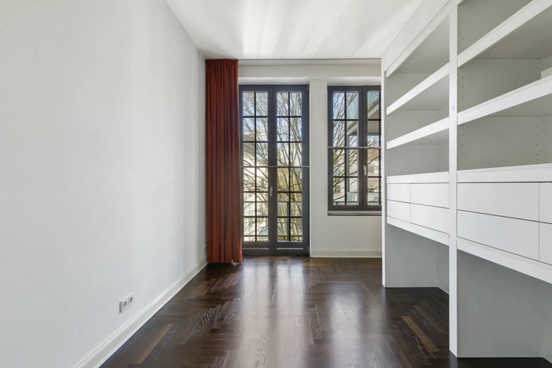 Heller Raum mit dunklem Holzboden, Regalwand und Fensterfront mit rotem Vorhang.