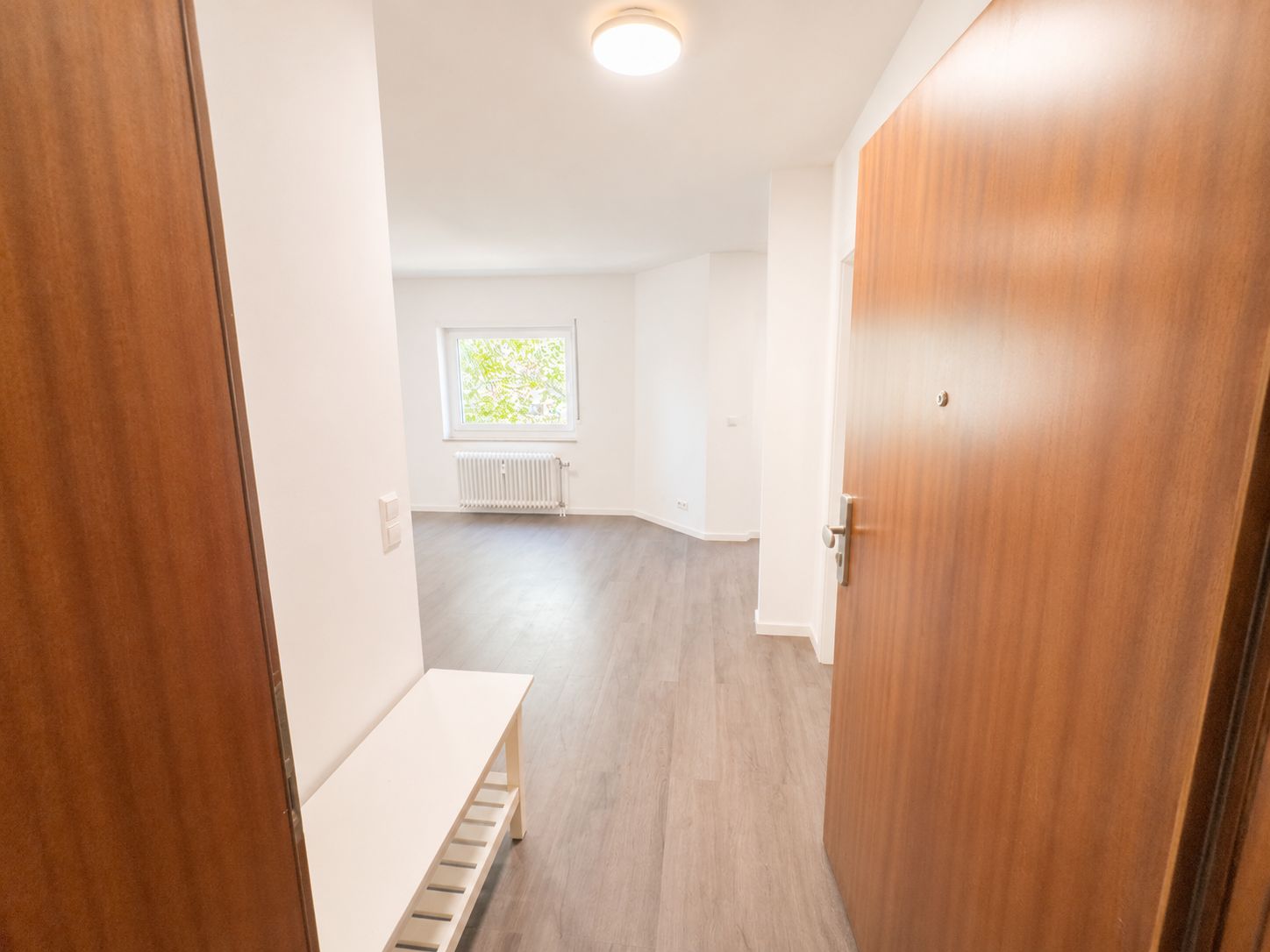 Offene Wohnungstür mit Blick auf einen hellen Raum, Fenster und Heizkörper, Boden in Holzoptik.