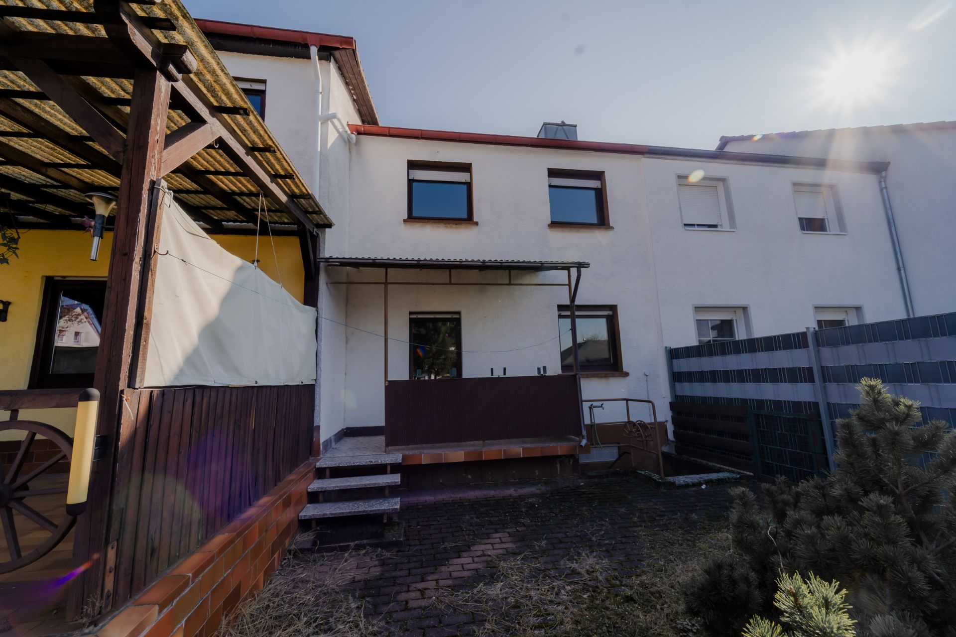 Rückseite eines weißen Wohnhauses mit Terrasse, Stufen und begrüntem Hof bei Sonnenschein.