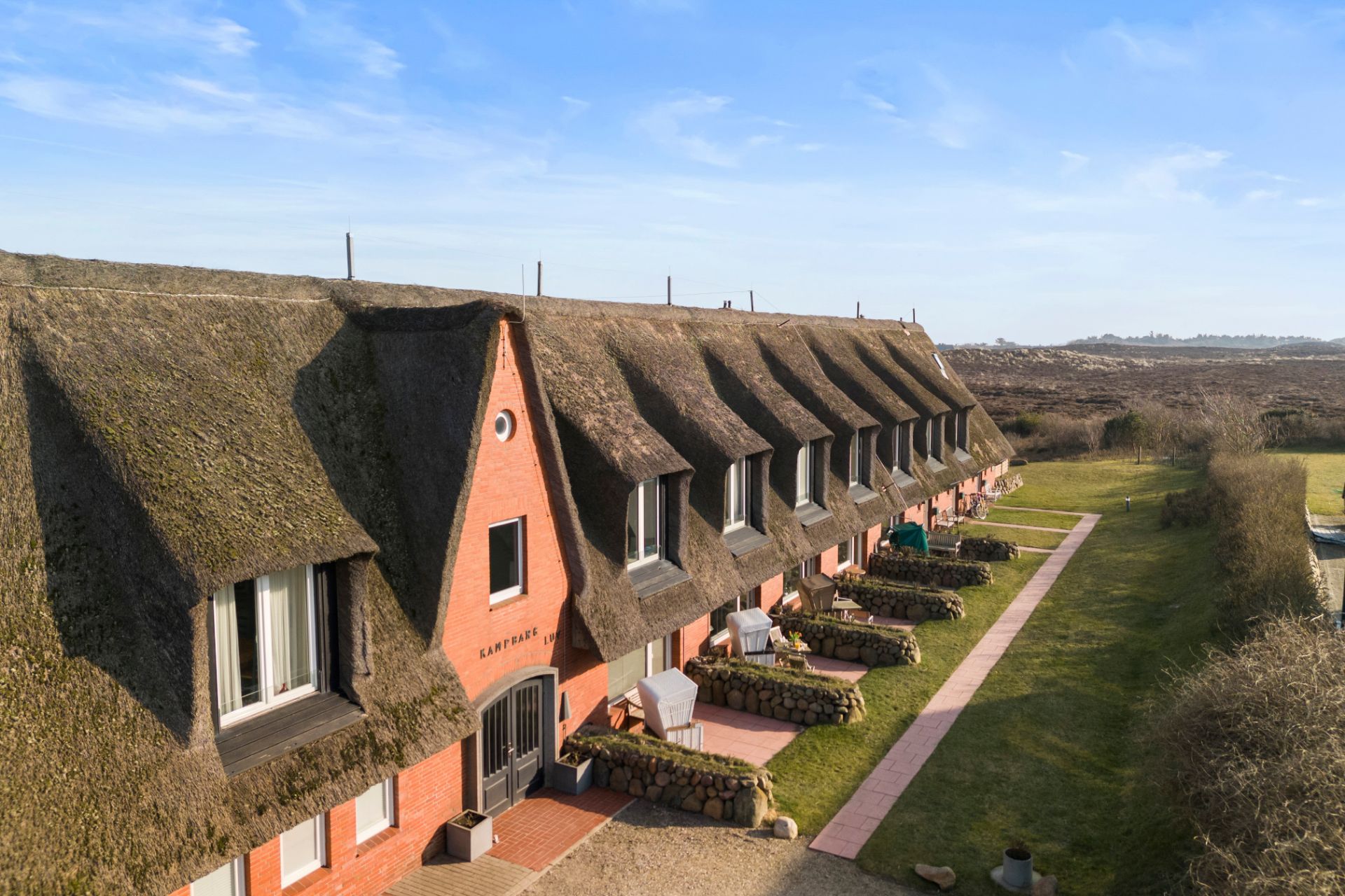 Reihenhaus mit Reetdach, roten Ziegelwänden und kleinen Terrassen im Grünen.