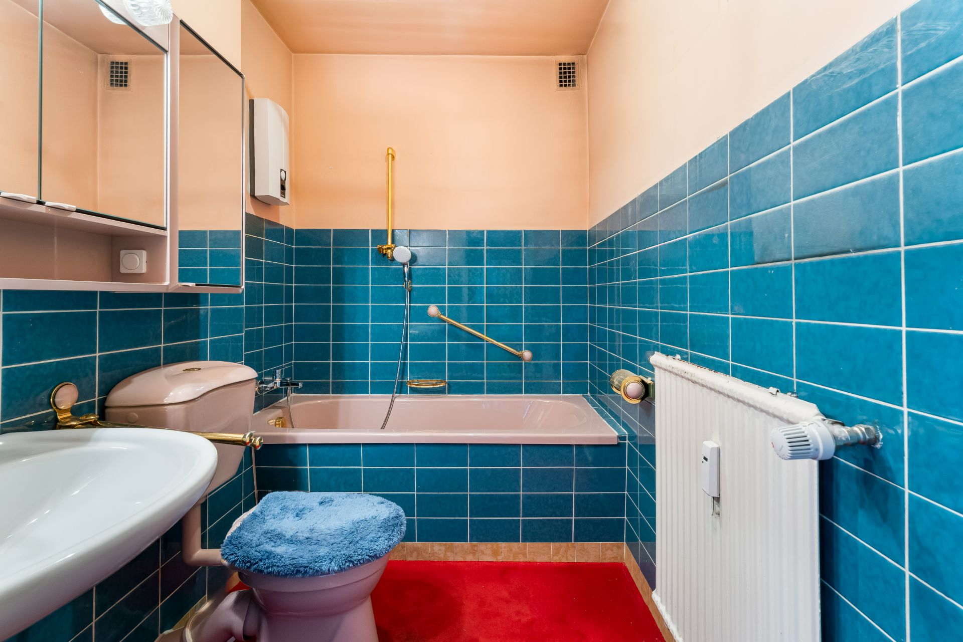 Badezimmer mit blauen Fliesen, Badewanne, Toilette mit blauem Bezug und rotem Teppich.