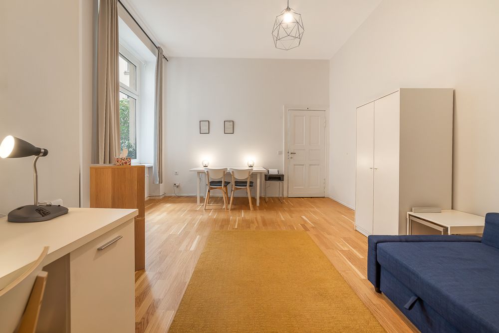 Helles Wohnzimmer mit Holzboden, Schreibtisch, Esstisch, blauem Sofa und weißem Schrank.