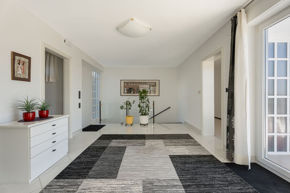 Heller Flur mit modernem Teppich, Kommode, Pflanzen und großen Fenstern.