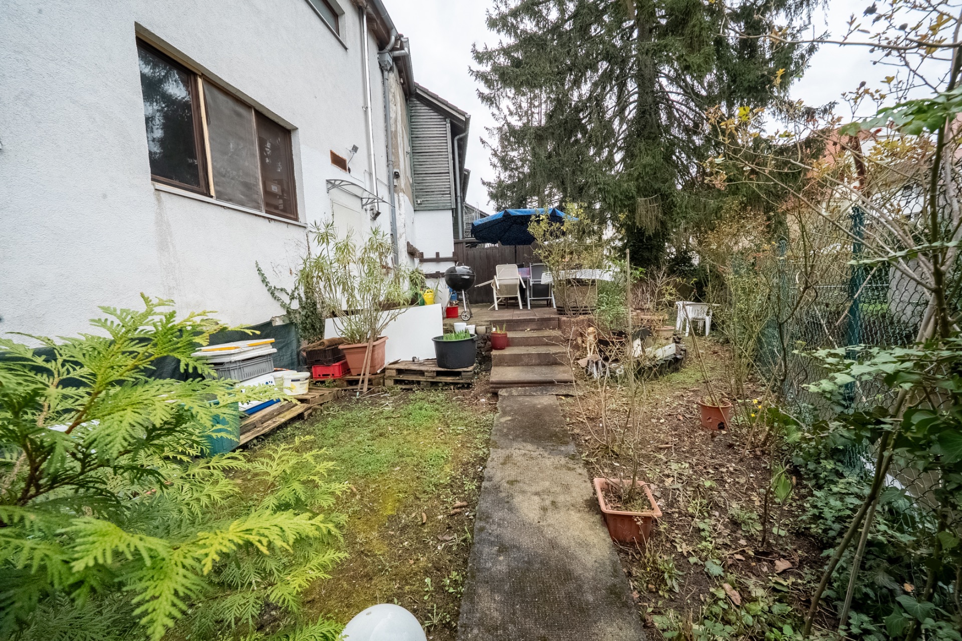Seitlicher Garten mit Terrasse, Stühlen, Pflanzenkübeln und einem Grill neben einem Wohnhaus.