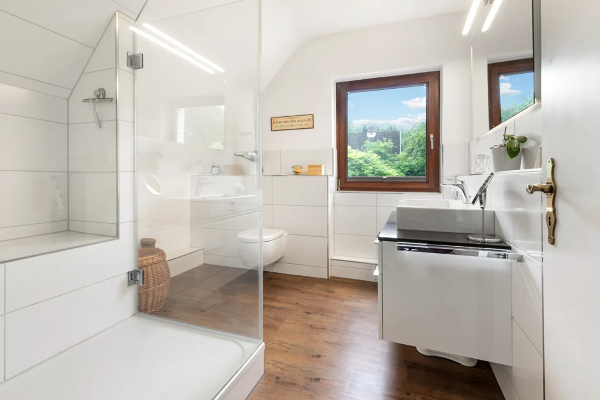 Modernes Badezimmer mit großer Dusche, Holzfußboden, Fenster und weißem Waschbecken.