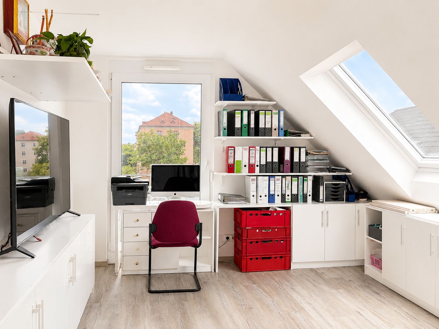 Helles Arbeitszimmer mit Schreibtisch, Computer, Regal mit Aktenordnern und großen Fenstern.