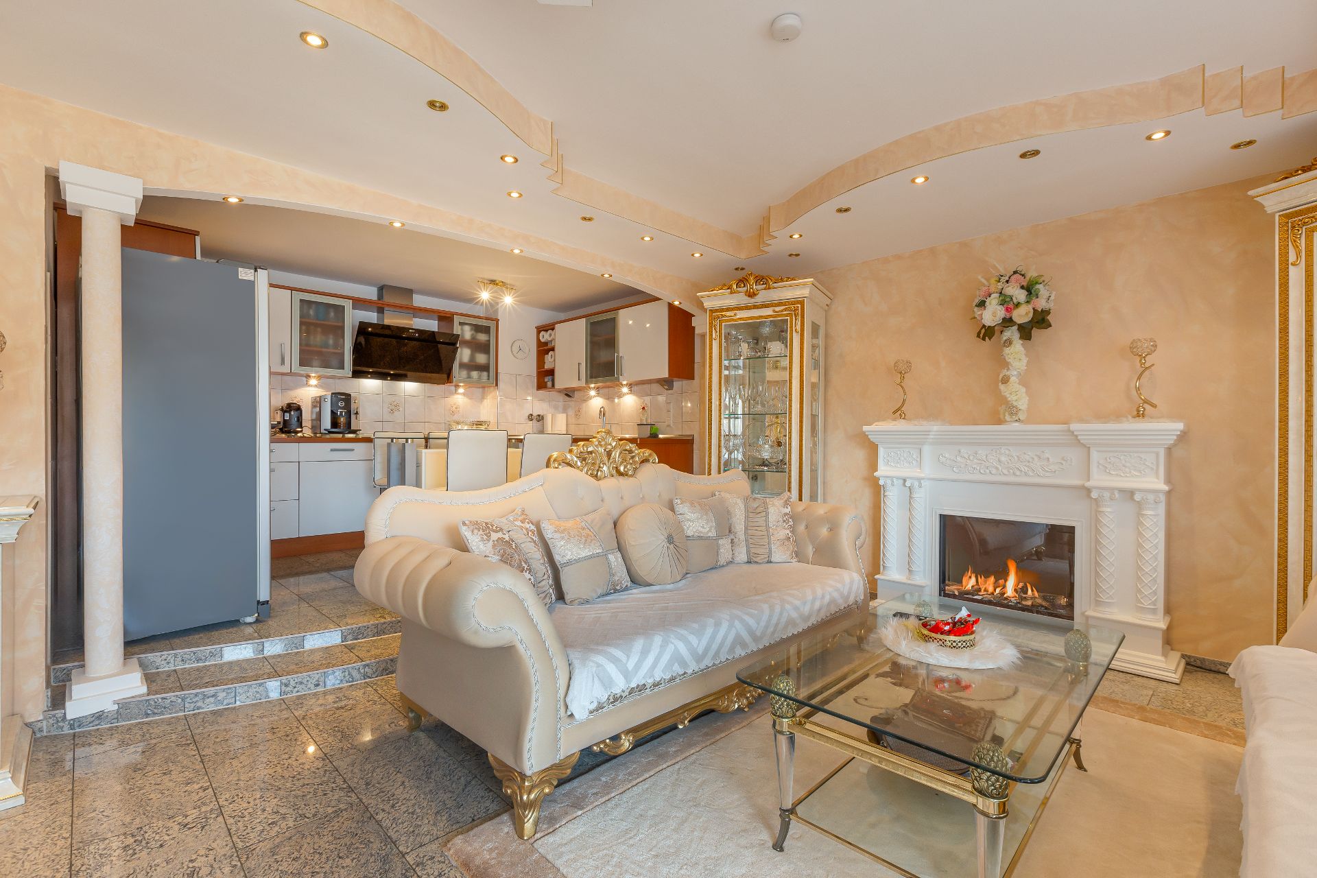 Elegantes Wohnzimmer mit Kamin, goldener Couch, Glas-Couchtisch und offener Küche im Hintergrund.