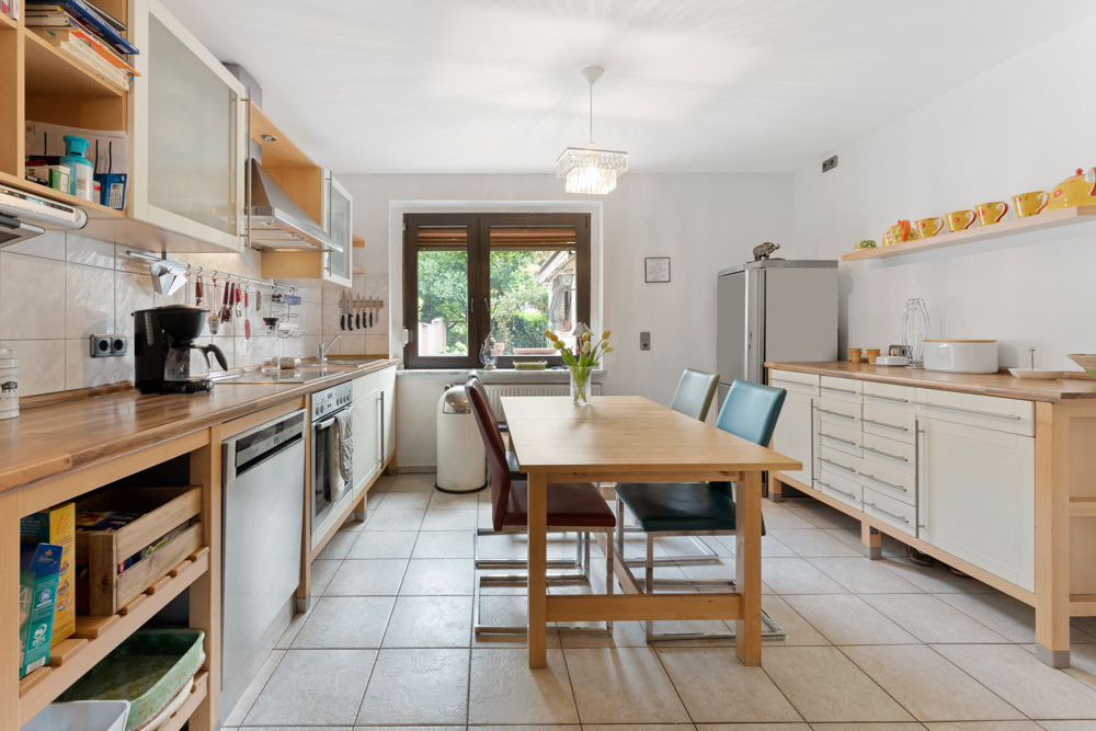 Moderne Küche mit Holztisch, vier Stühlen, großen Fenstern und Küchengeräten auf Fliesenboden.