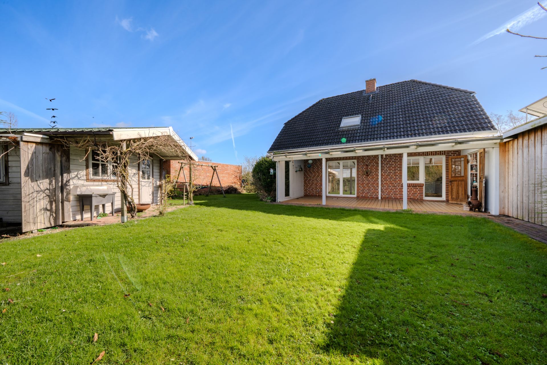 Großer Garten mit Rasen, Terrasse, Haus und kleinem Nebengebäude bei sonnigem Wetter.
