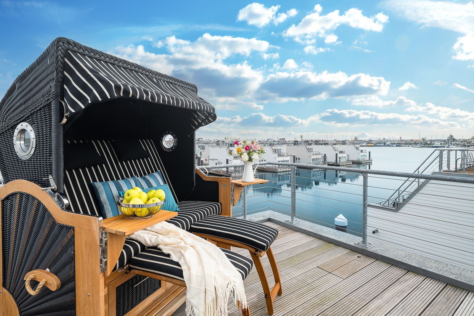 Strandkorb auf einer Terrasse mit Blick auf einen Hafen, daneben ein Tisch mit Blumen und eine Schale mit Äpfeln.