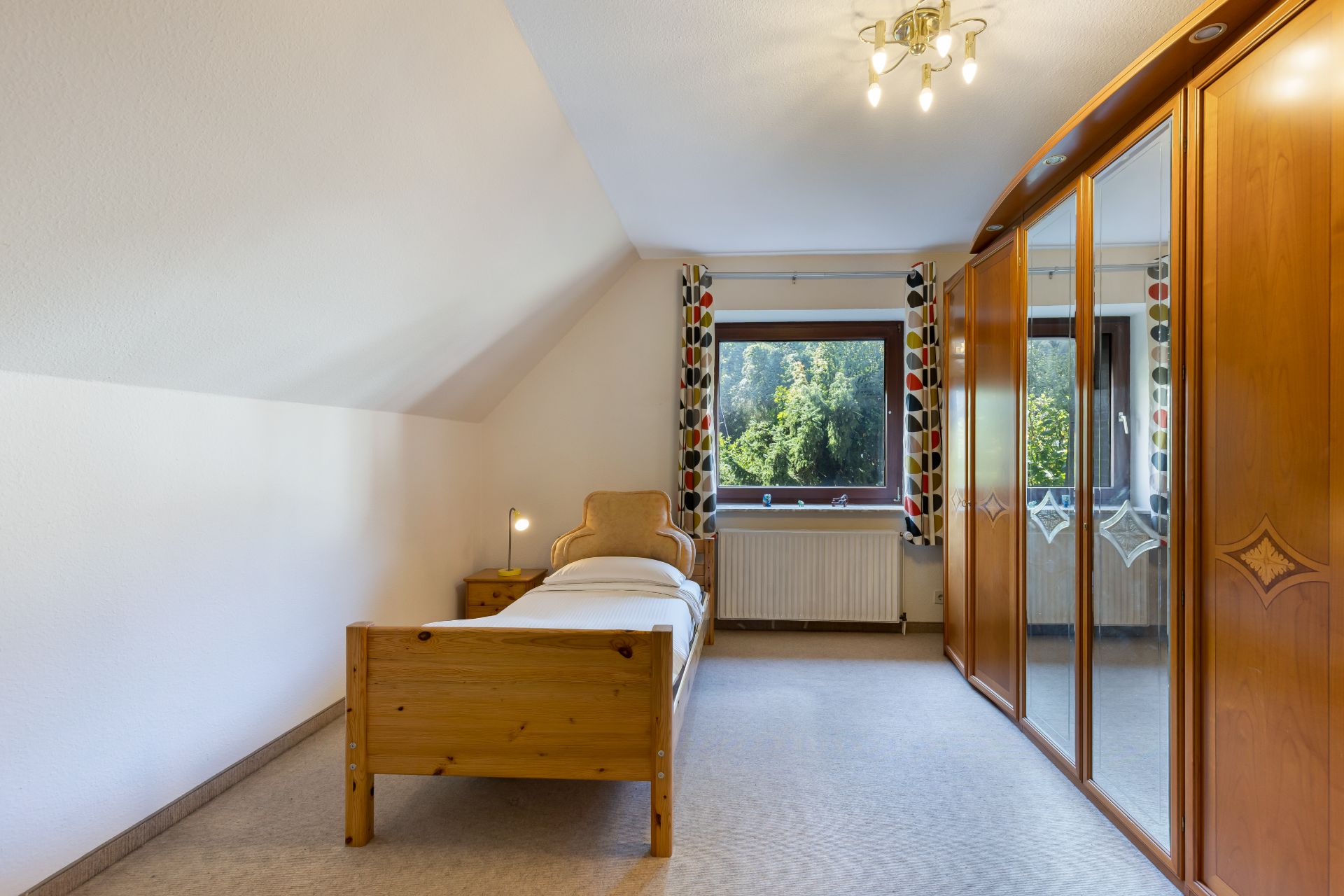 Helles Schlafzimmer mit Einzelbett, Nachttisch, großem Holzkleiderschrank und Fenster mit Blick ins Grüne.