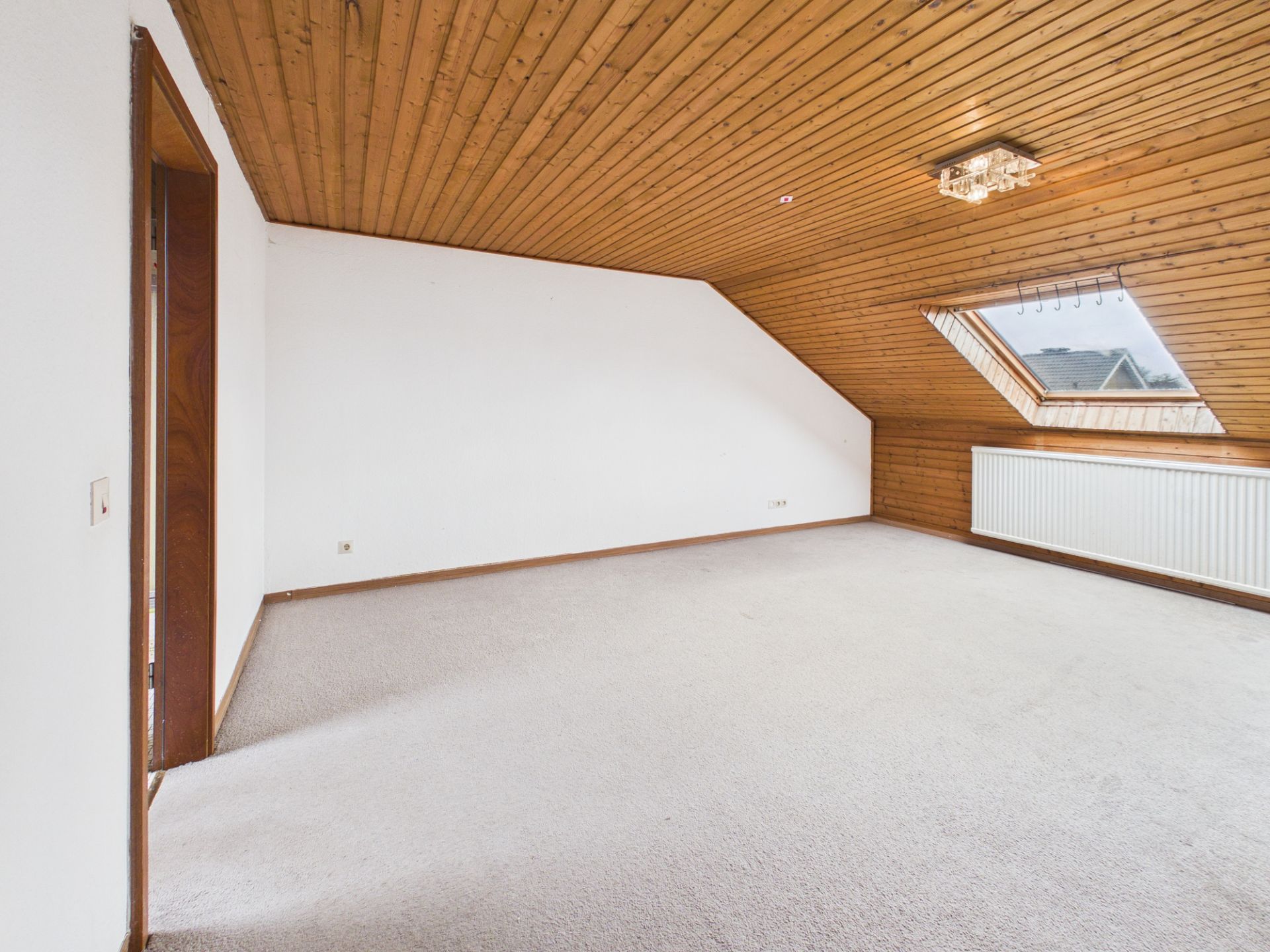 Leeres Zimmer mit schräger Holzdecke, großem Fenster und Heizkörper unter der Fensterbank.