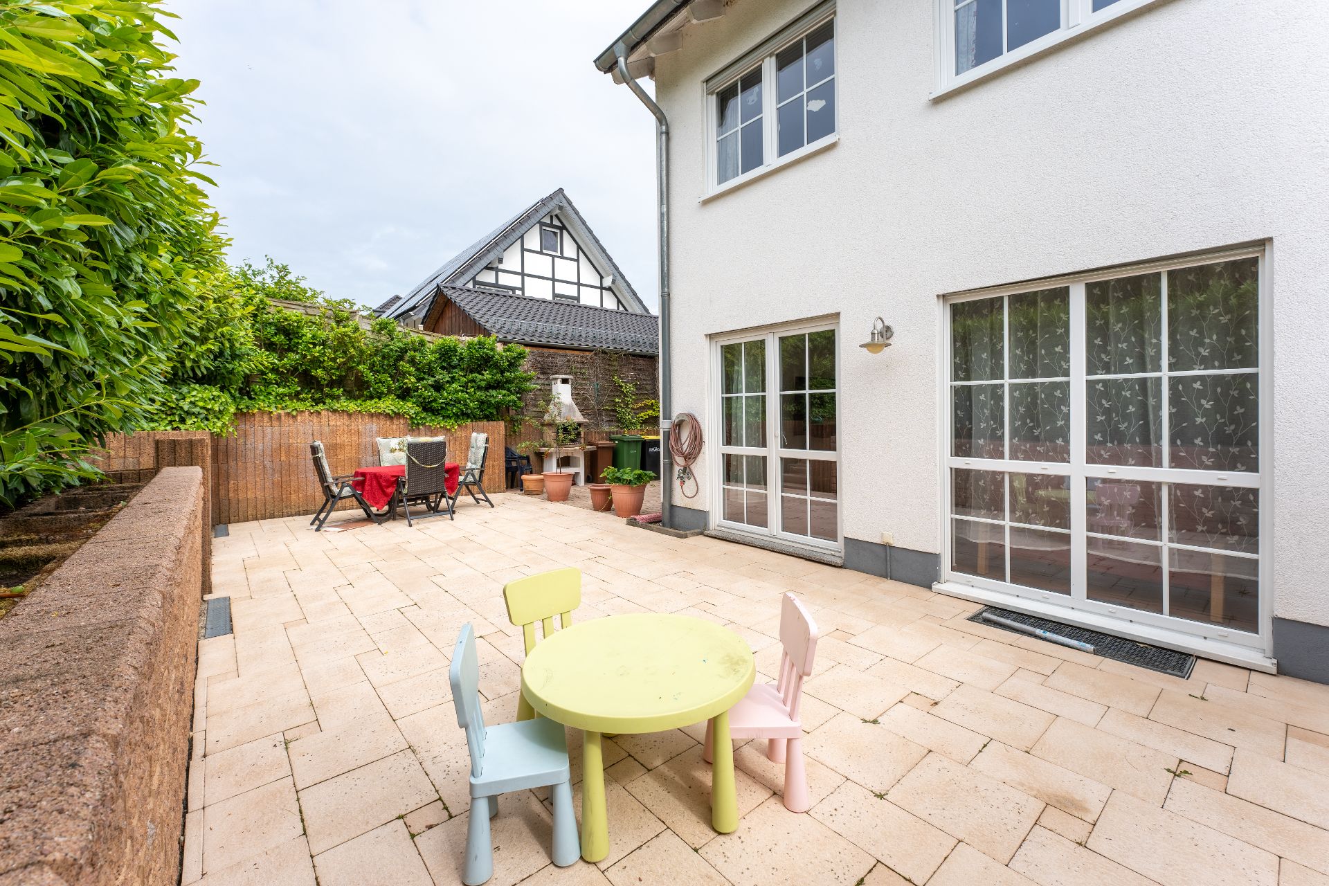 Große Terrasse mit Kinder- und Gartentisch, umgeben von Pflanzen und einem weißen Haus.