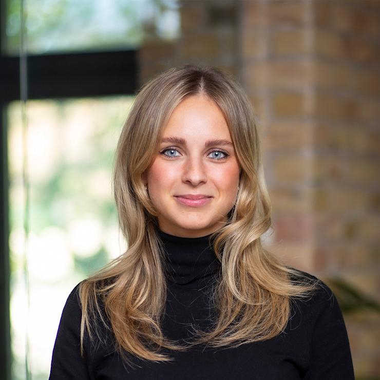 Junge Frau mit blondem Haar und gestreiftem Blazer steht mit verschränkten Armen im Innenraum.