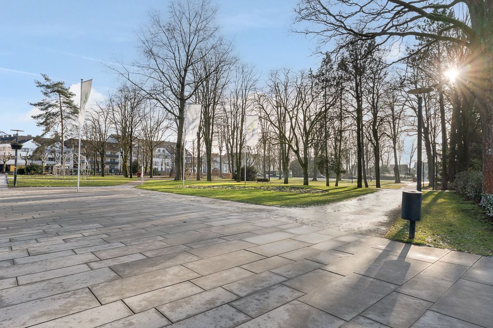 Gepflasterter Platz mit Rasenflächen, Bäumen und weißen Fahnen bei sonnigem Wetter.