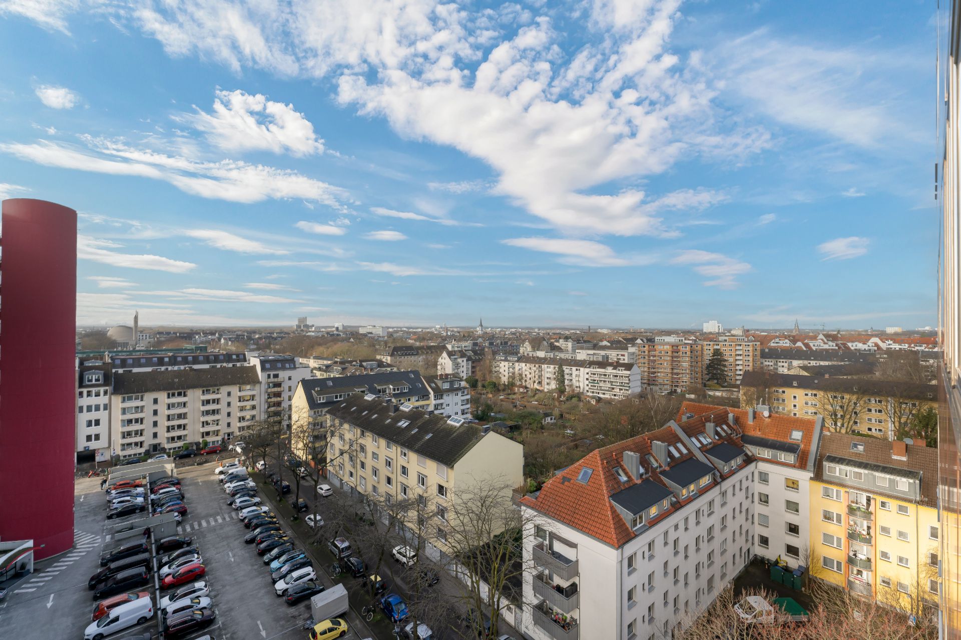 Blick aus der Höhe auf Wohngebäude, parkende Autos und einen blauen Himmel mit Wolken.
