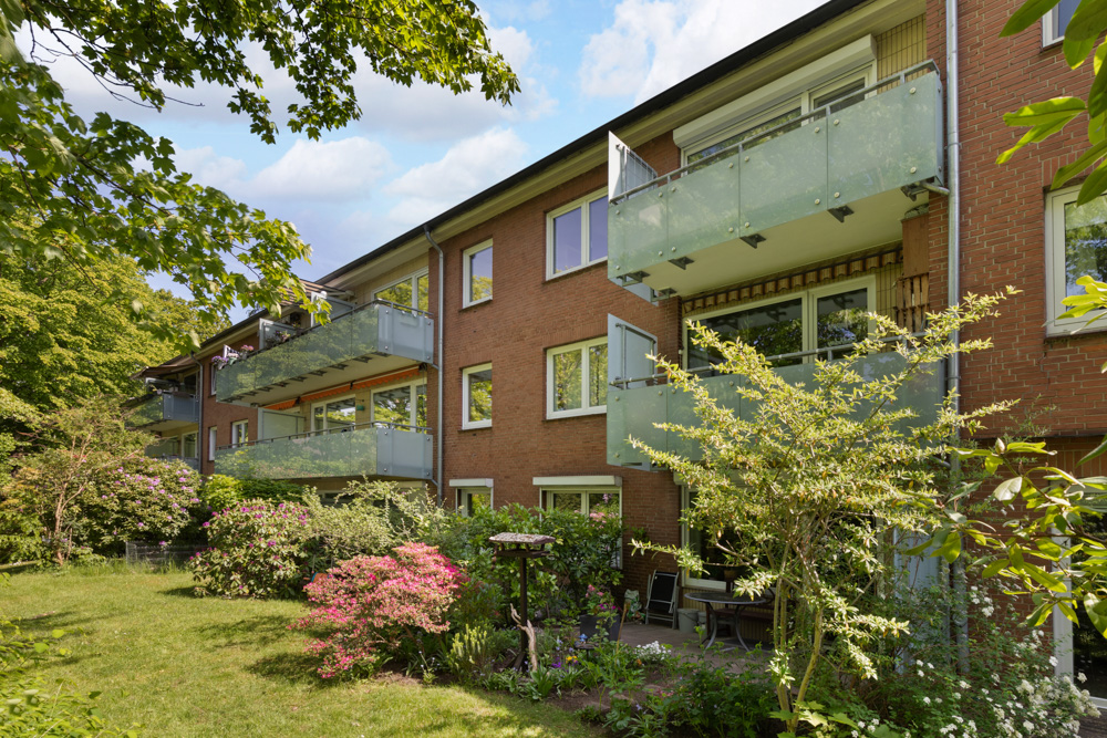 Mehrfamilienhaus mit Glasbalkonen und Garten mit Blumen und Sträuchern.