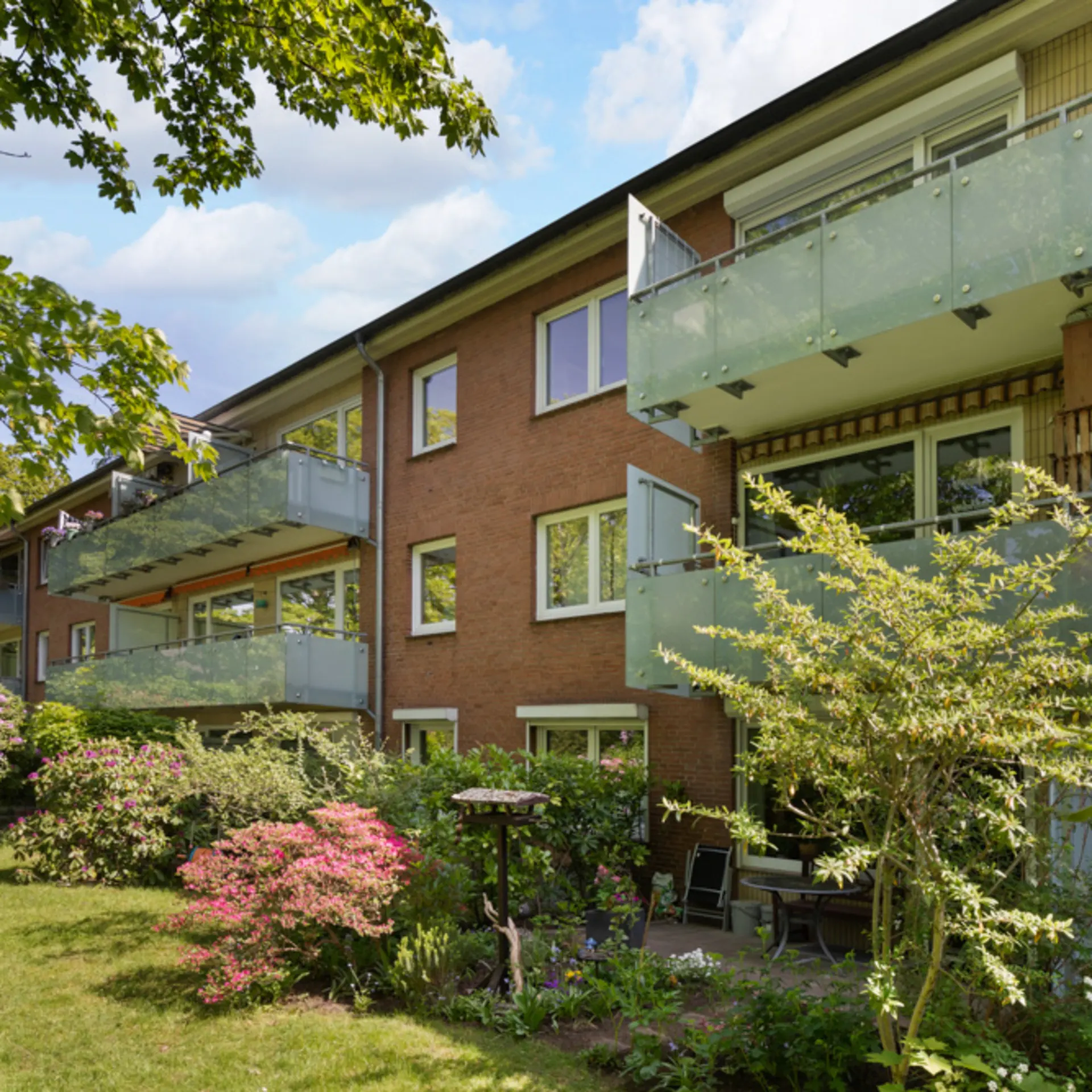 Mehrfamilienhaus mit Glasbalkonen und Garten mit Blumen und Sträuchern.