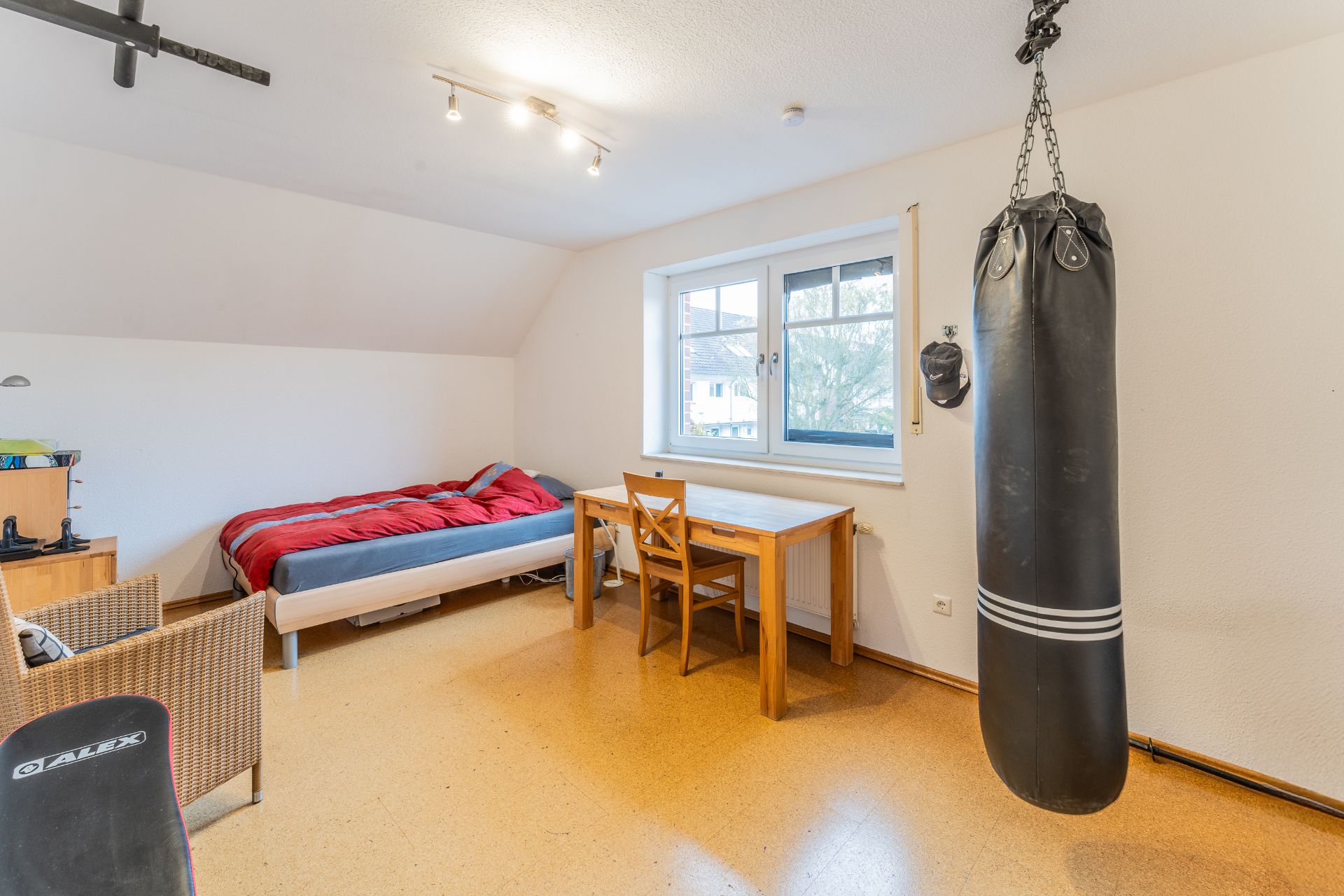 Helles Zimmer mit Bett, Schreibtisch, Stuhl und einem Boxsack an der Decke.