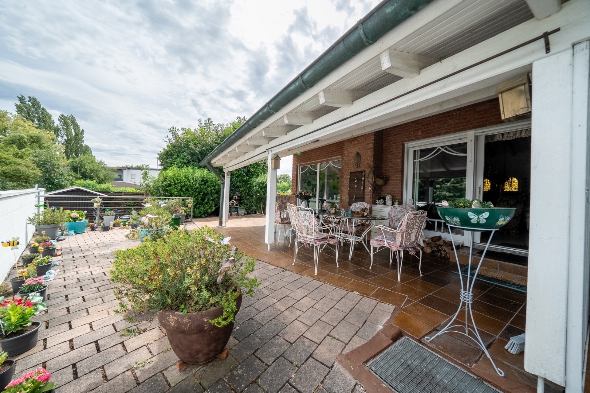 Überdachte Terrasse mit Gartentisch, Stühlen und vielen Blumenkübeln neben einem Wohnhaus.