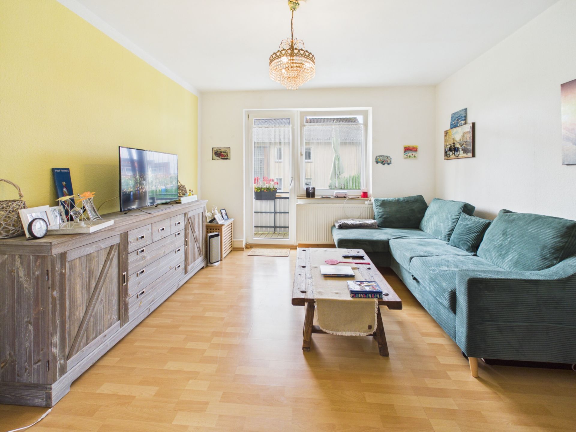 Helles Wohnzimmer mit grünem Ecksofa, Holztisch, Sideboard und TV, gelber Wand und großem Fenster.