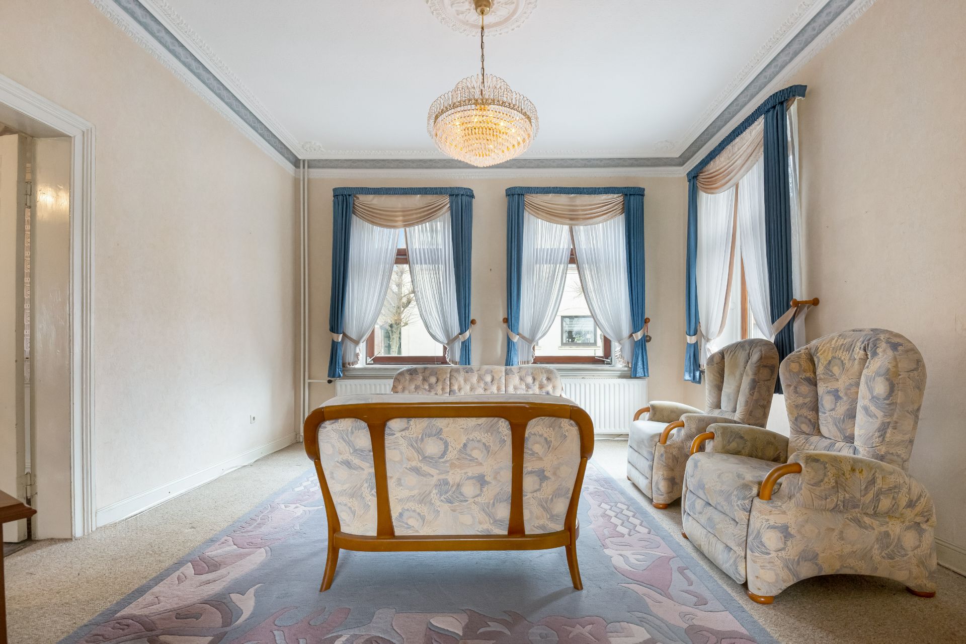 Wohnzimmer mit gemustertem Sofa, zwei Sesseln, Kronleuchter und drei Fenstern mit blauen Vorhängen.