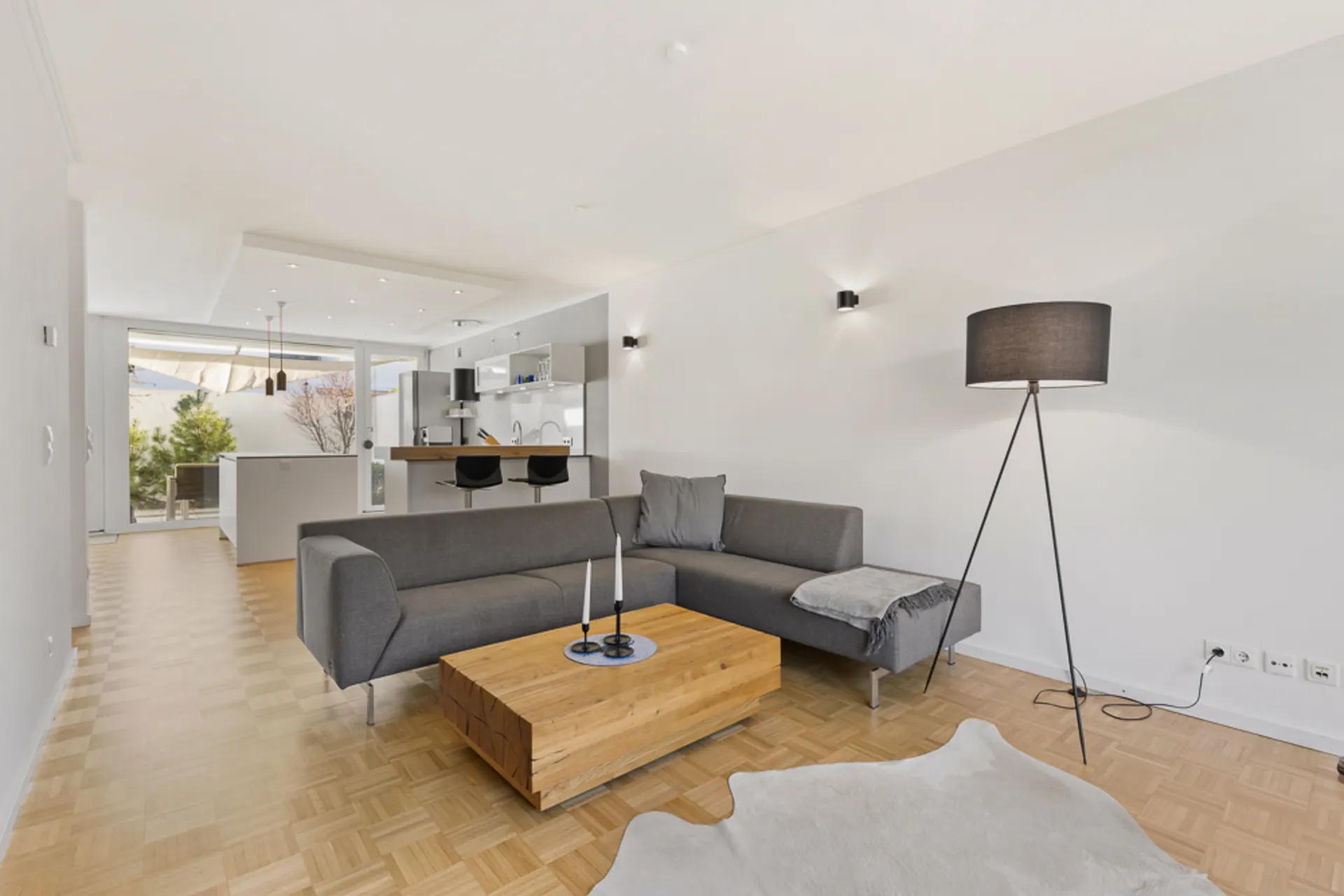 Modernes Wohnzimmer mit grauem Sofa, Holz-Couchtisch, Stehlampe und offener Küche im Hintergrund.
